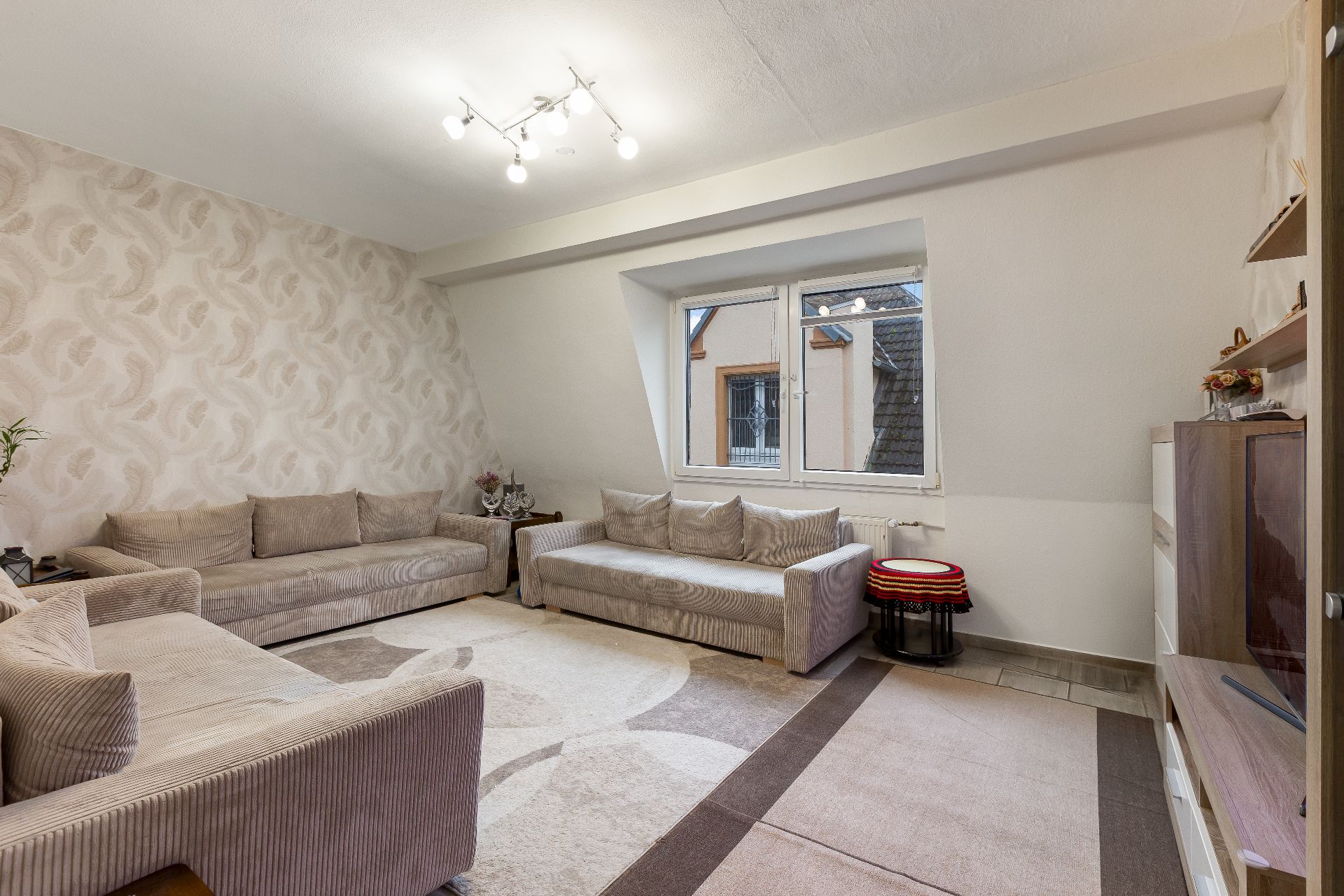 Wohnzimmer mit zwei beigen Sofas, Teppich, Fenster und Deckenbeleuchtung.