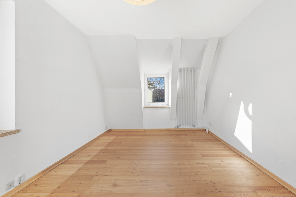 Heller leerer Raum mit Holzfußboden, weiß gestrichenen Wänden und einem Fenster mittig.