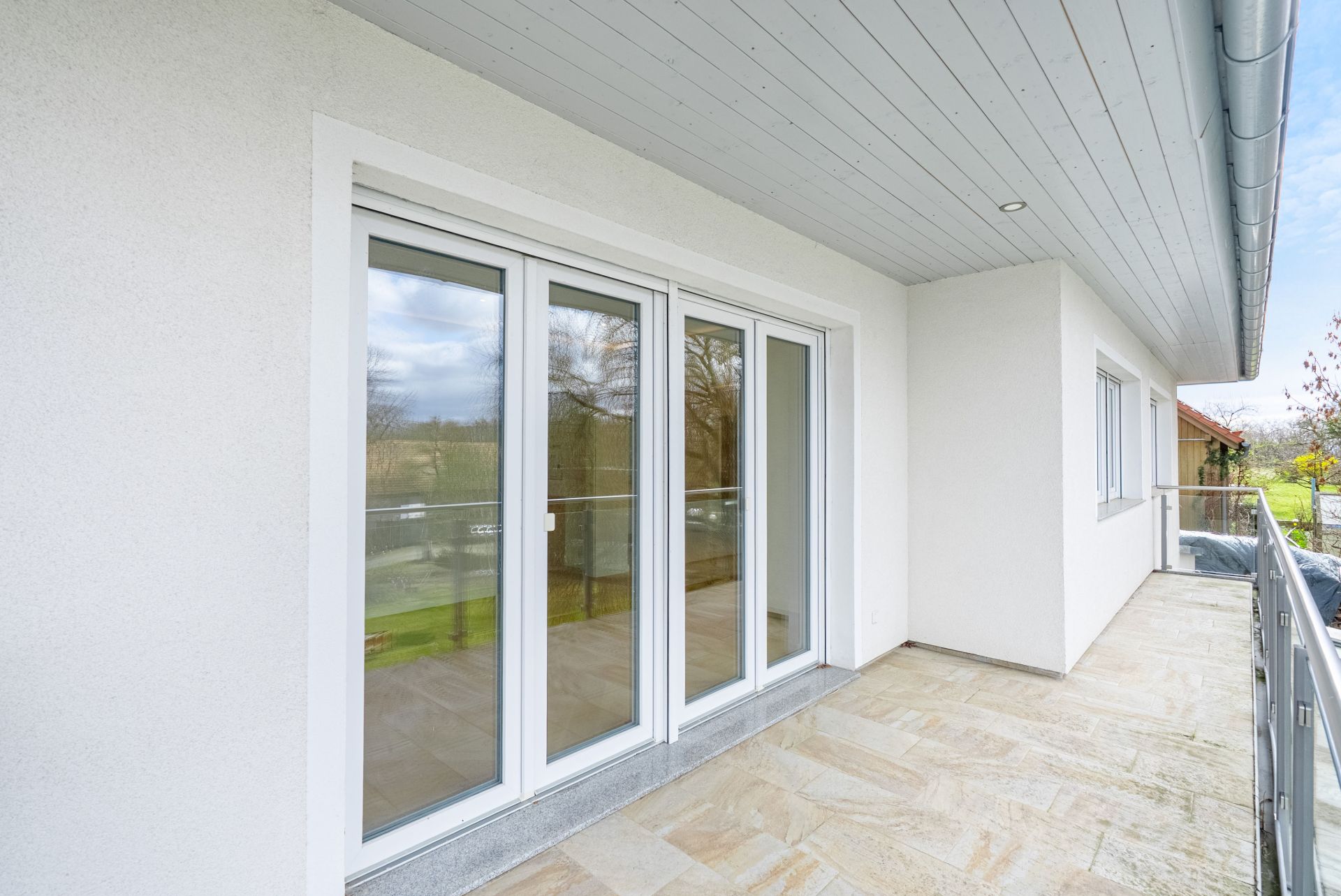 Helle Terrasse mit Glasgeländer und großen weißen Fenstertüren an einem Wohnhaus.