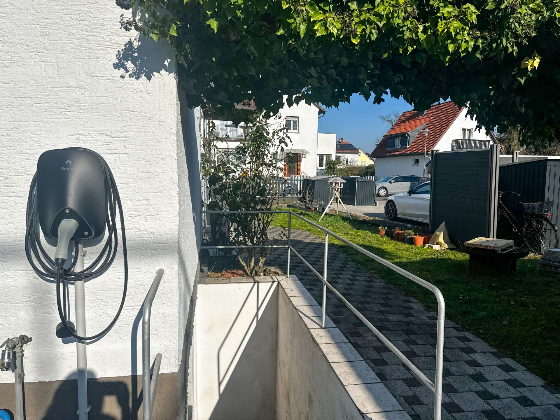 Ladestation an einer weißen Hauswand, gepflasterter Weg und Garten, Autos und Fahrräder im Hintergrund.