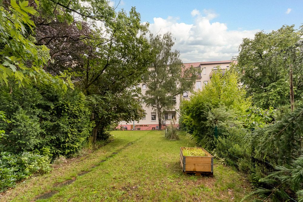 Grüner Gartengrund mit Bäumen, Sträuchern und einem Hochbeet; Wohnhaus im Hintergrund.