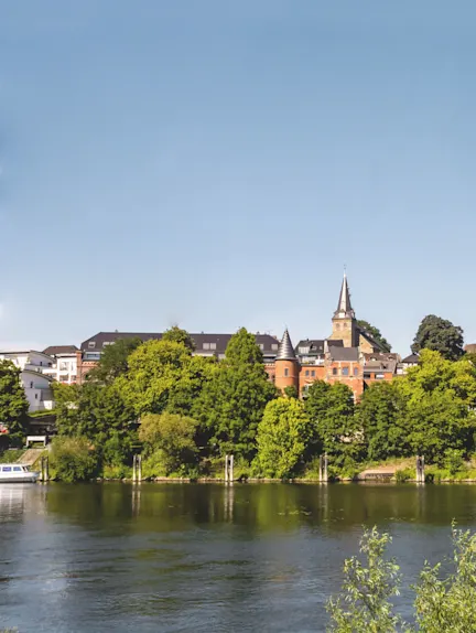Häuser und Bäume am Ufer eines Flusses, im Hintergrund ein Turm mit spitzem Dach.