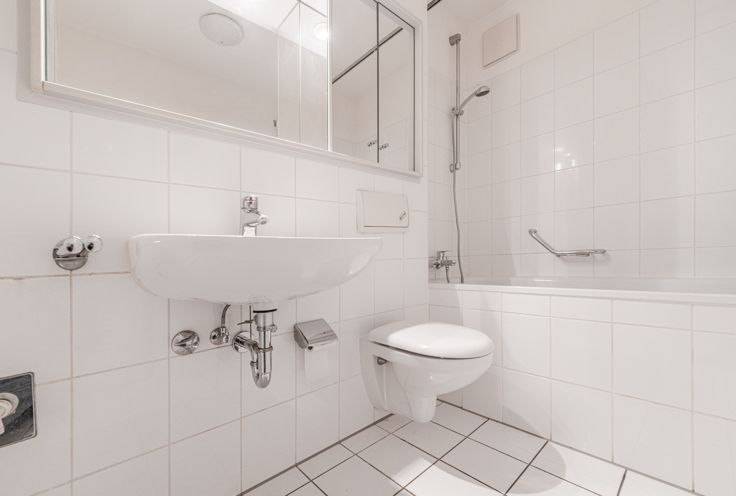 Helles Badezimmer mit Wandwaschbecken, Toilette und Badewanne mit Duschkopf.