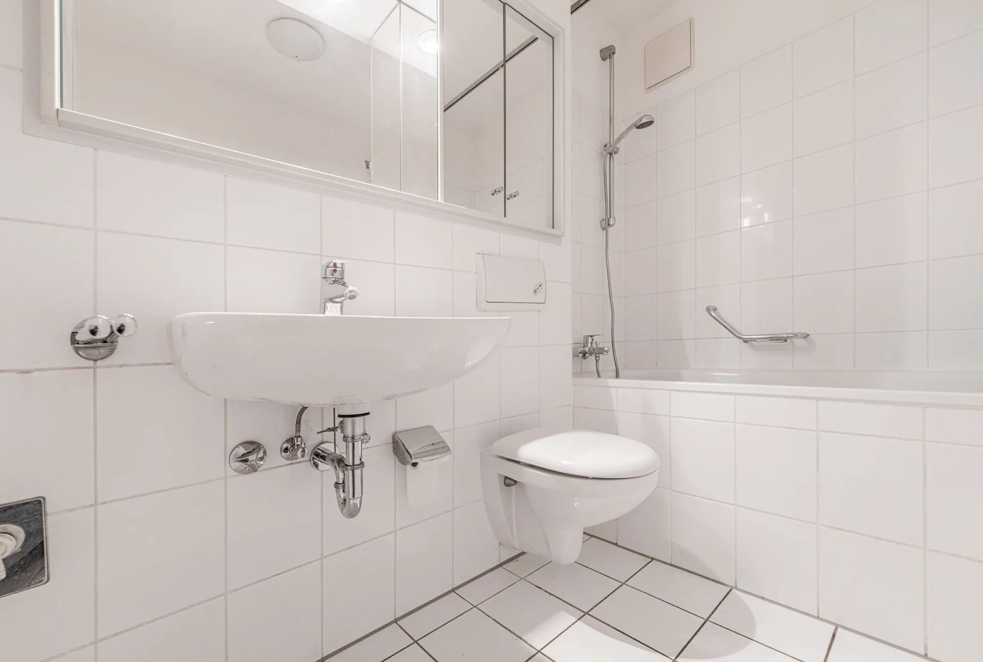 Helles Badezimmer mit Wandwaschbecken, Toilette und Badewanne mit Duschkopf.