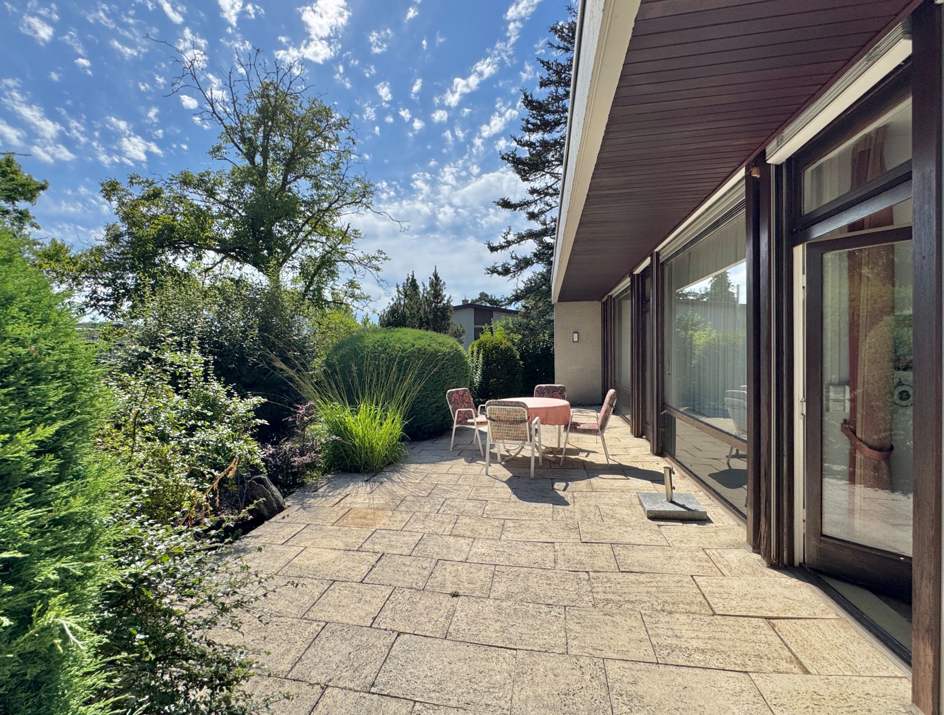 Sonnige Terrasse mit Gartentisch, Stühlen und umliegenden Pflanzen an einem Wohnhaus.