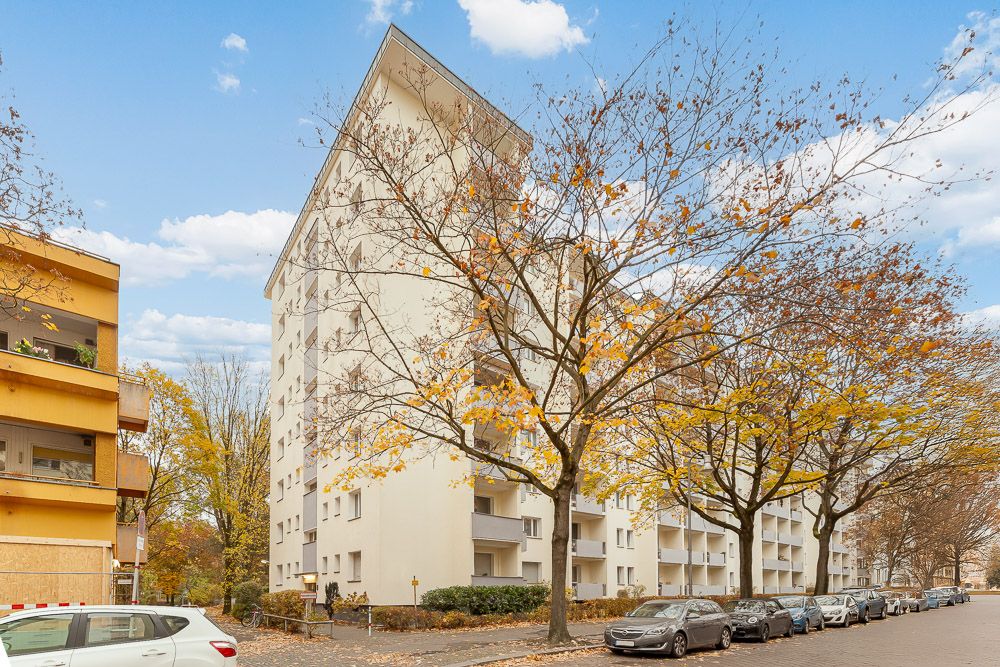 Mehrstöckiges Wohnhaus mit Balkonen an einer Baumallee, Autos parken am Straßenrand.