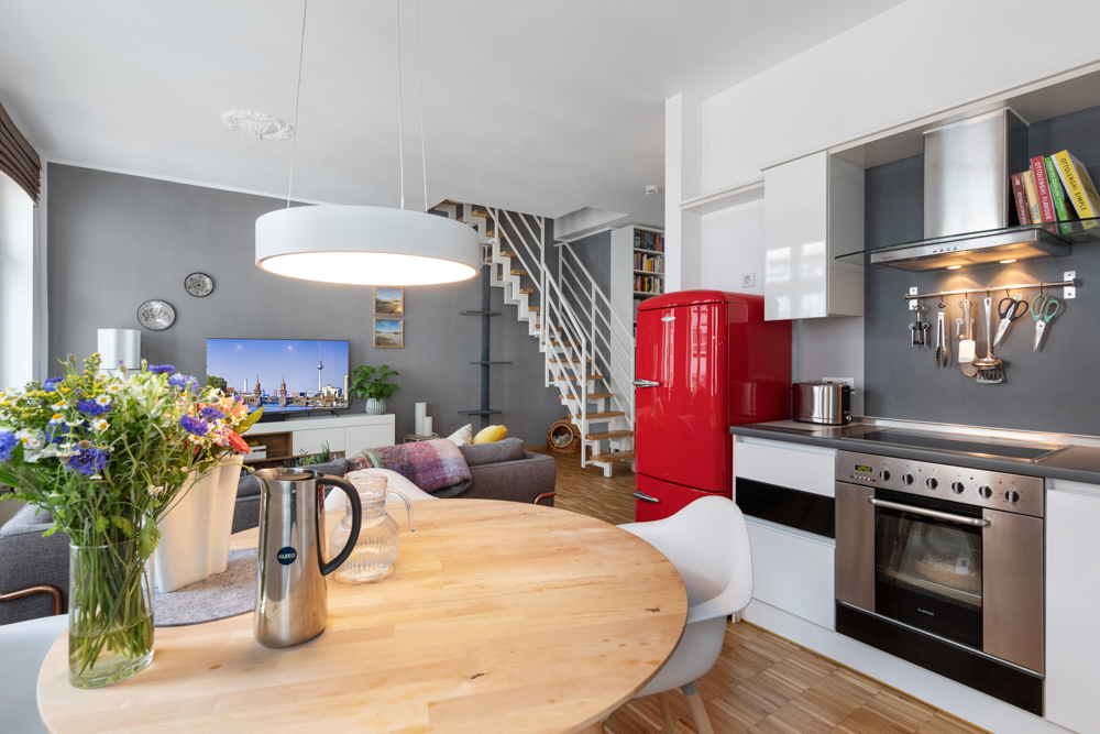 Moderne Küche und Wohnzimmer mit rotem Kühlschrank, Holztisch und Treppe im Hintergrund.