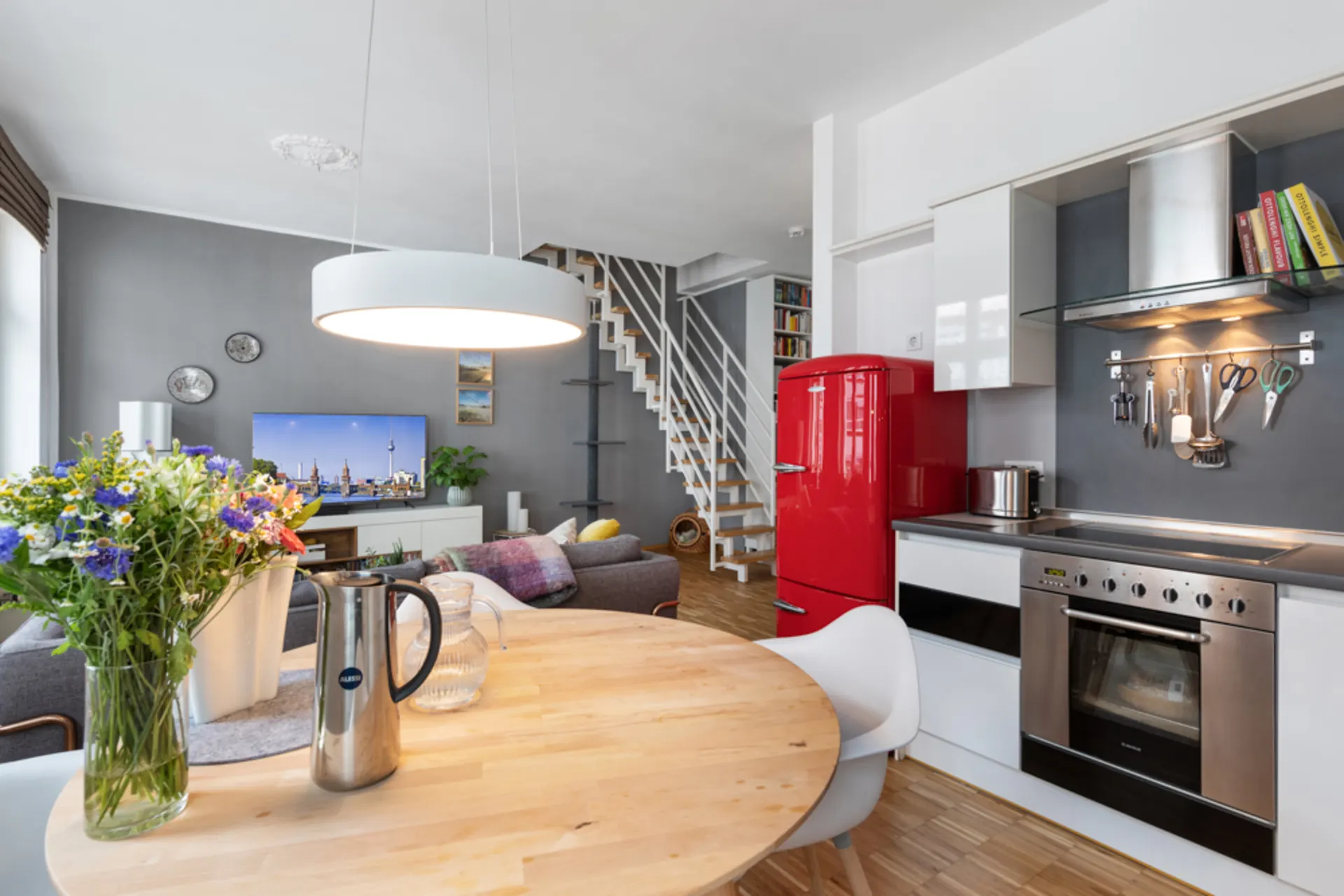 Moderne Küche und Wohnzimmer mit rotem Kühlschrank, Holztisch und Treppe im Hintergrund.