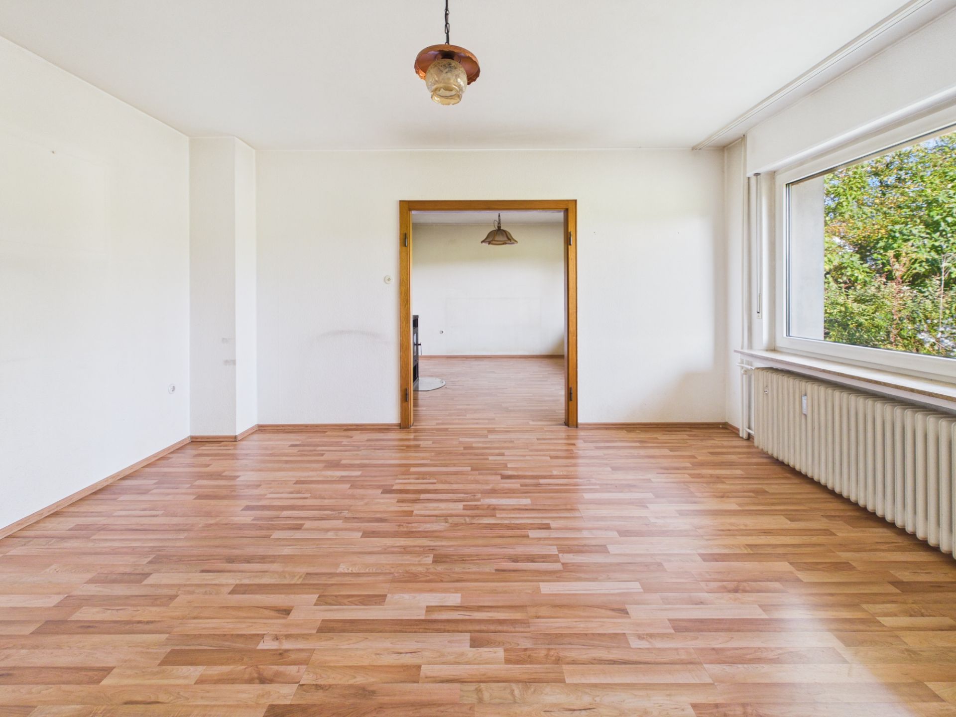 Leerer Raum mit Holzfußboden, weißer Wand, großem Fenster und Blick in einen angrenzenden Raum.