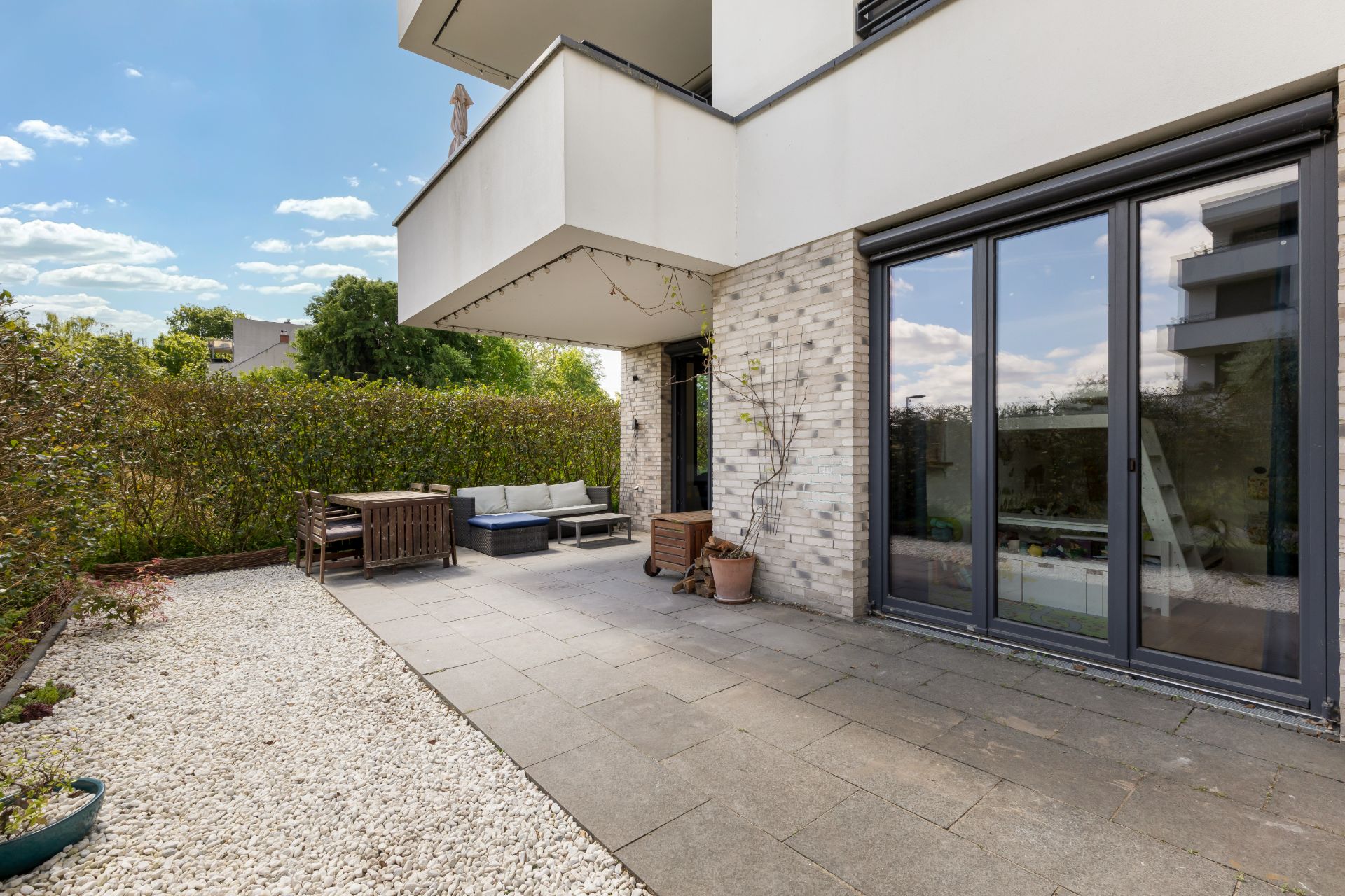 Moderne Terrasse mit Sitzbereich, großen Fenstern und Kiesstreifen neben einer Hecke.