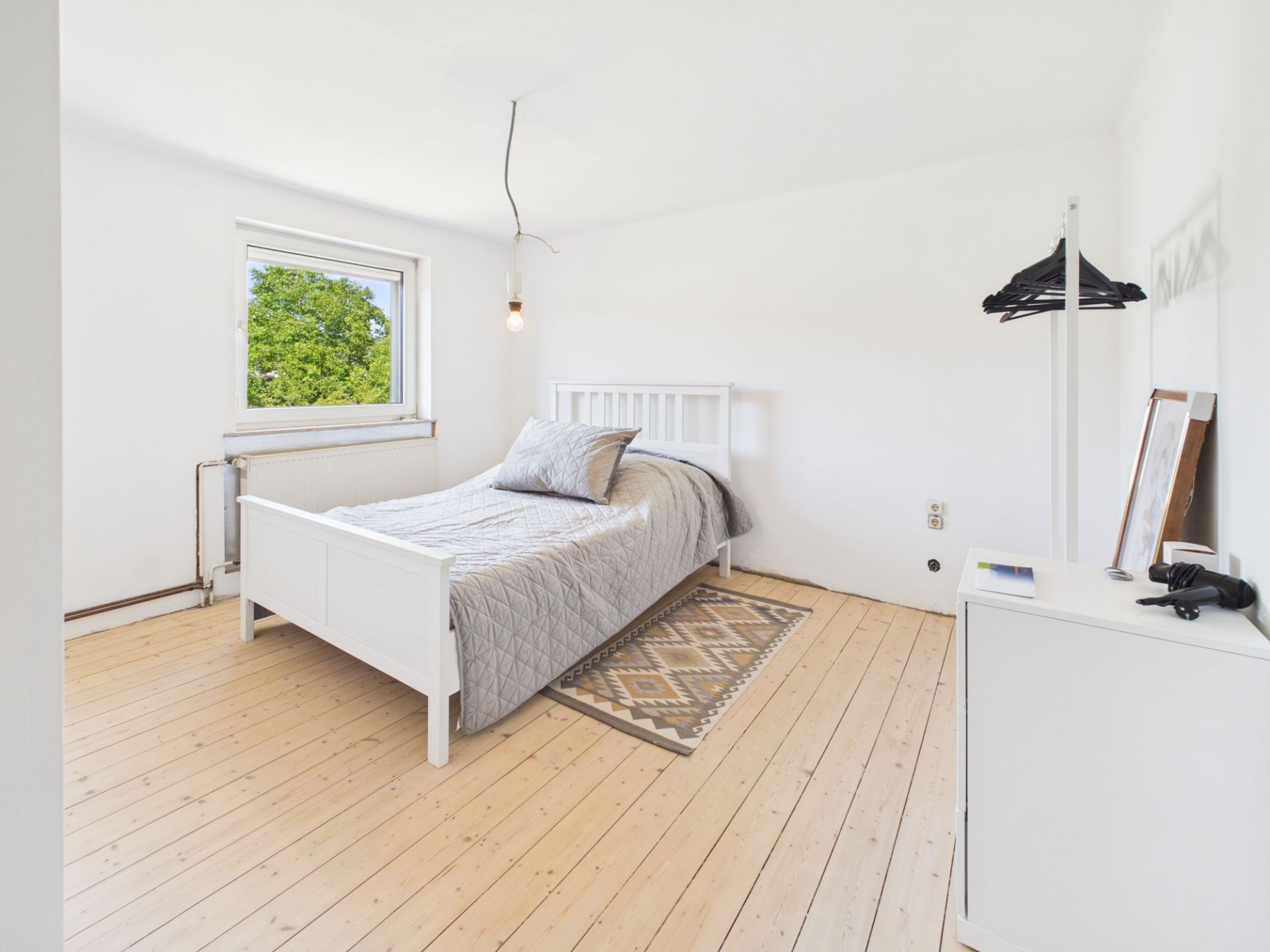 Helles Schlafzimmer mit Einzelbett, Teppich, Fenster und Kommode auf Holzboden.