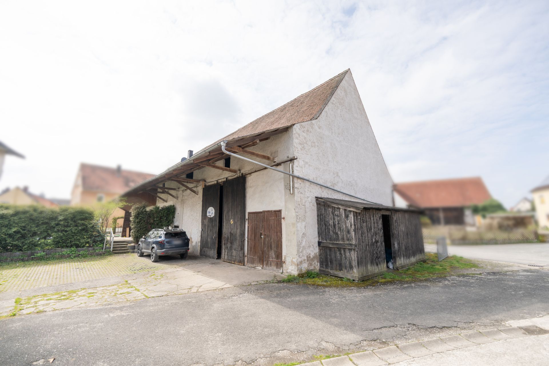 Altes, weißes Gebäude mit Satteldach, großem Holztor und kleinem Anbau, Auto davor geparkt.