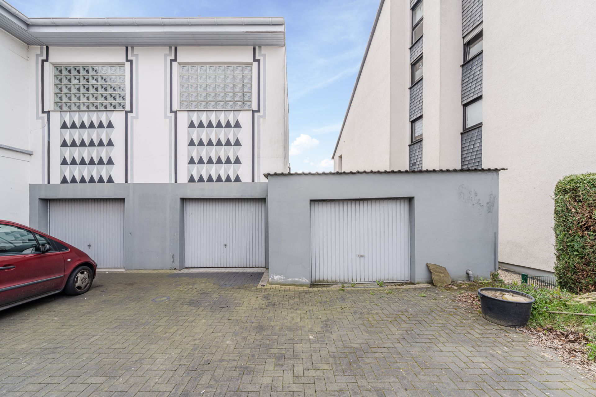 Zwei graue Garagentore vor einem Wohnhaus mit geometrischen Mustern, links steht ein rotes Auto.