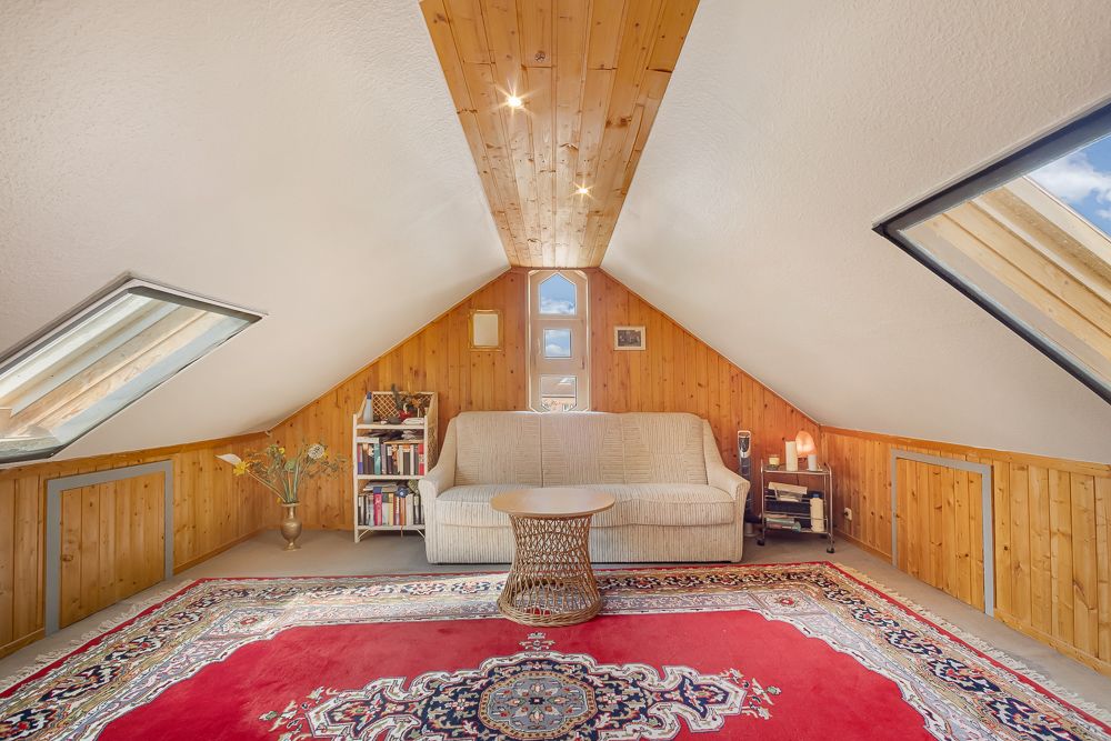 Gemütlicher Dachraum mit Sofa, Teppich, Dachfenstern, Holzverkleidung und Regalen.