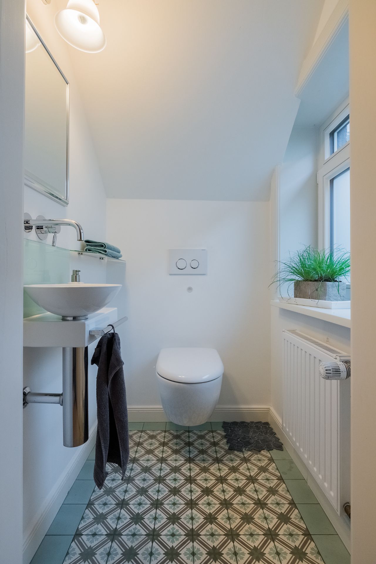 Kleines Badezimmer mit Wandtoilette, Waschbecken, Spiegel und Fenster mit Pflanze.