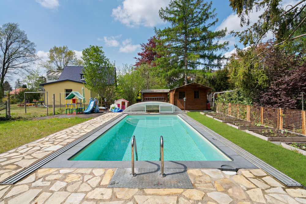 Garten mit rechteckigem Pool, Spielplatz, Haus und Holzhütte im Hintergrund.