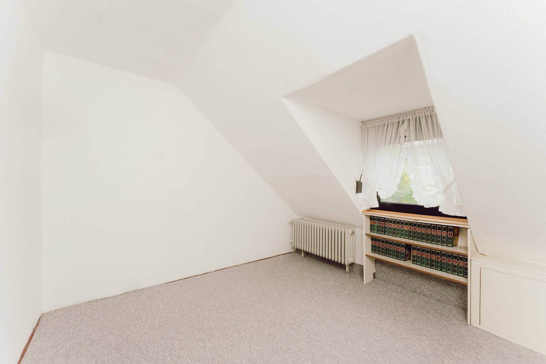 Heller Dachraum mit Teppichboden, Heizkörper und Regal unter schrägem Fenster.