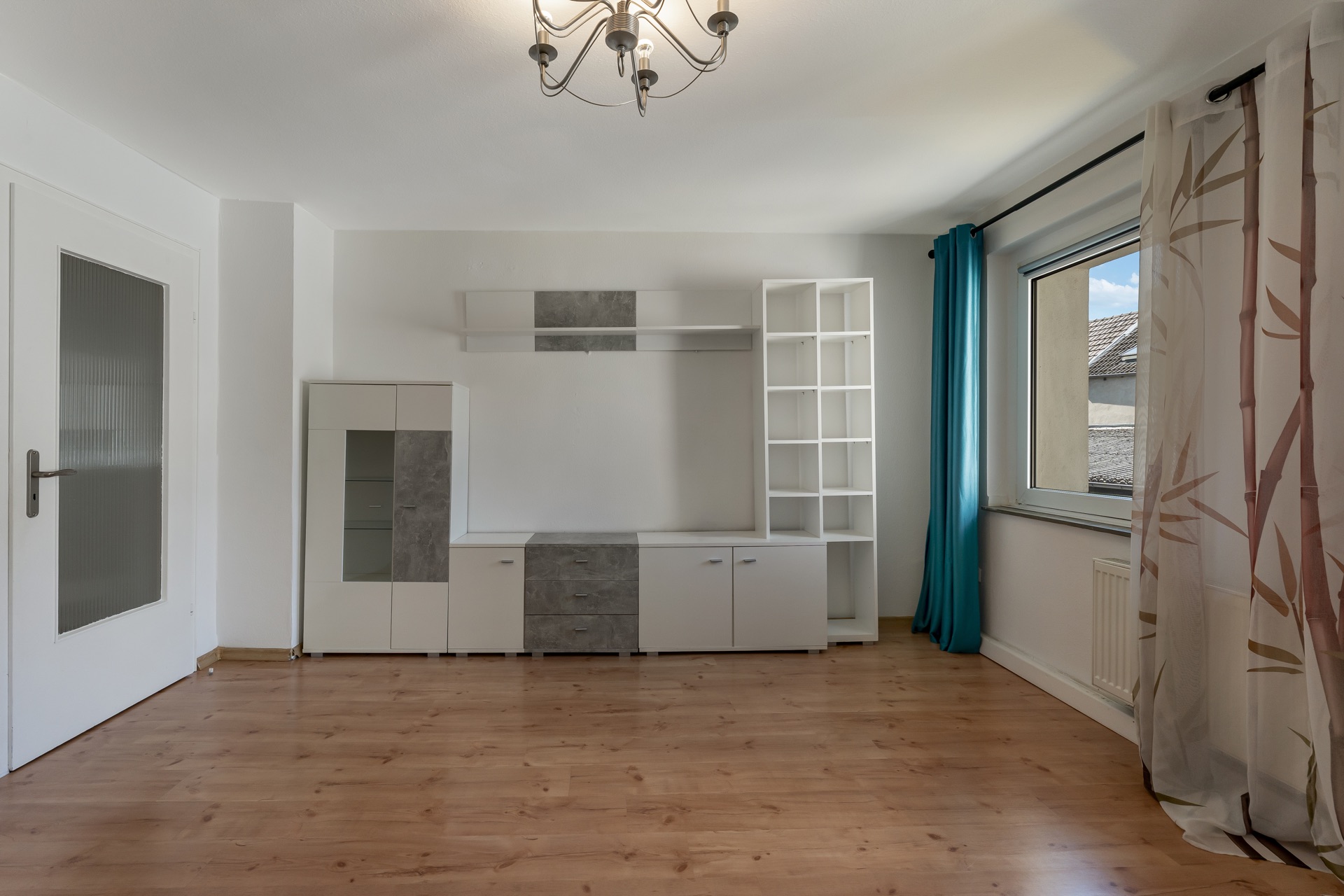 Wohnzimmer mit Holzfußboden, weißem Schrank, großem Fenster und Vorhängen.