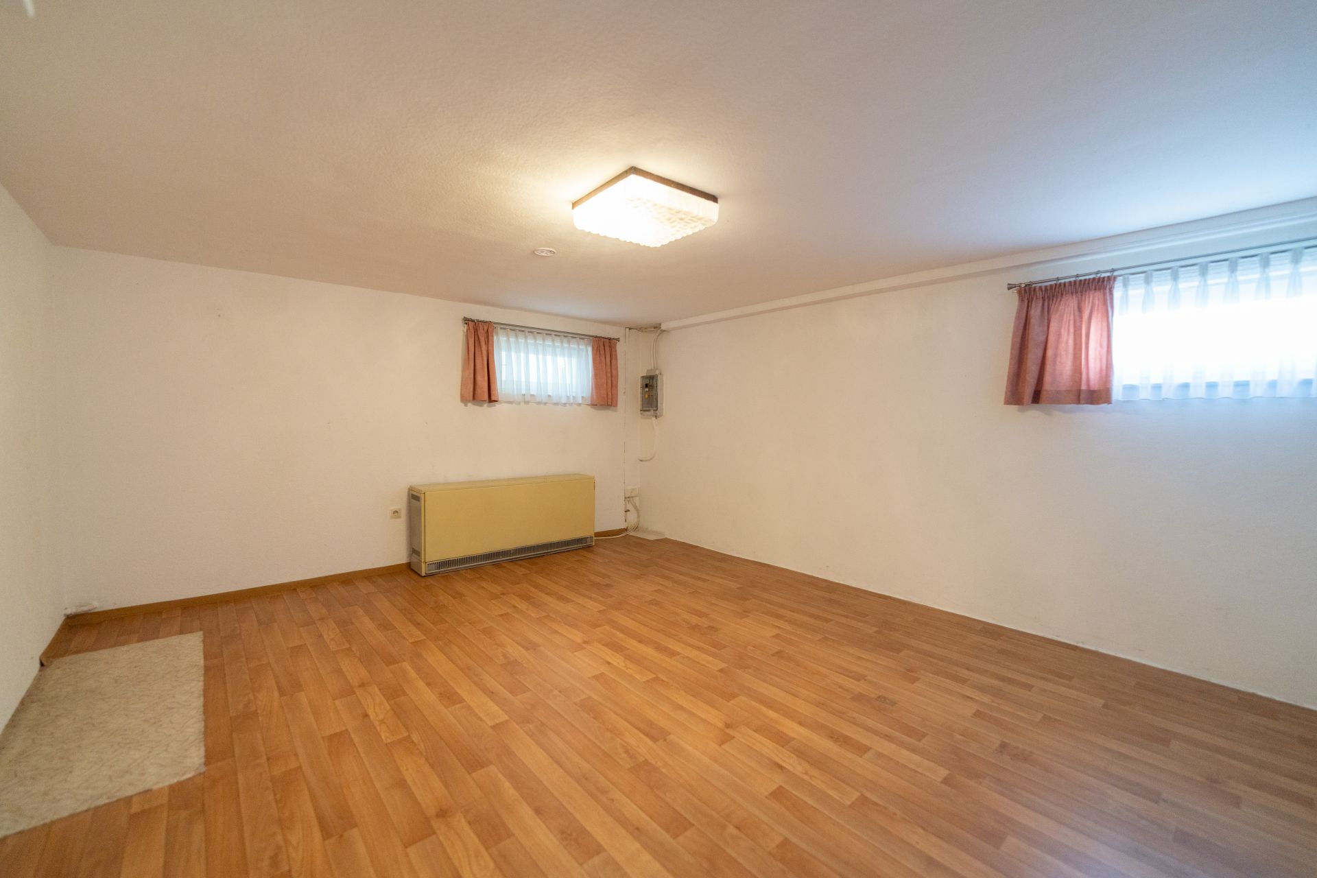 Leerer Raum mit Holzfußboden, weißen Wänden, zwei Fenstern mit Vorhängen und Deckenleuchte.