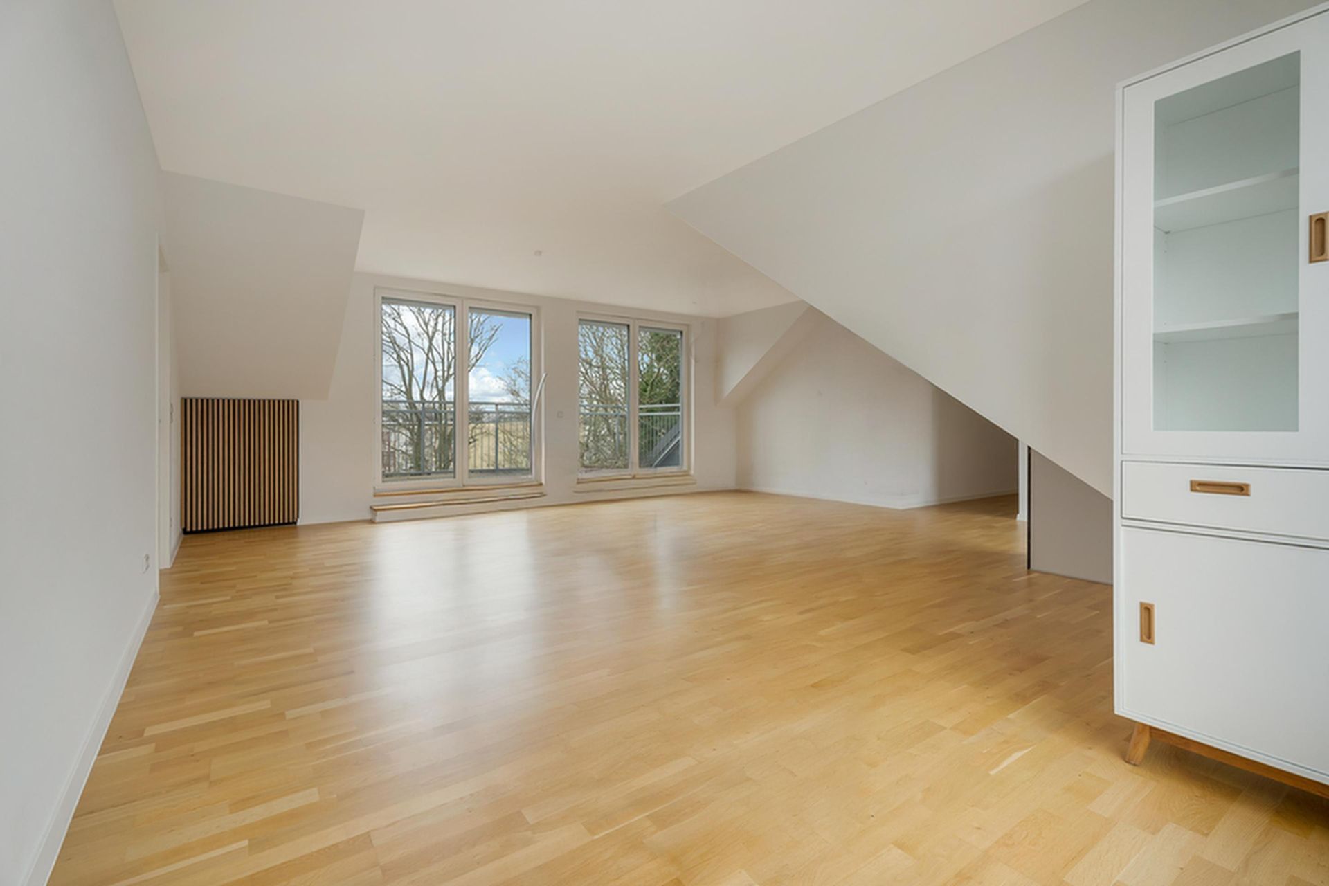 Helles Wohnzimmer mit Parkettboden, großen Fenstern und einer weißen Vitrine.