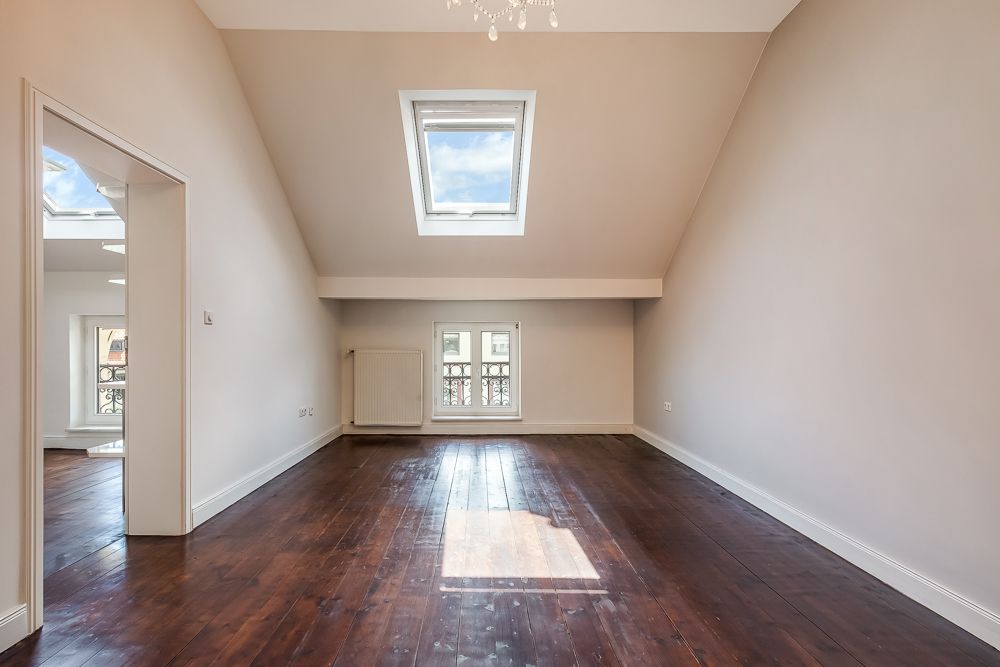 Leerer Raum mit dunklem Holzboden, Dachfenster und kleinem Fenster an der Stirnseite.