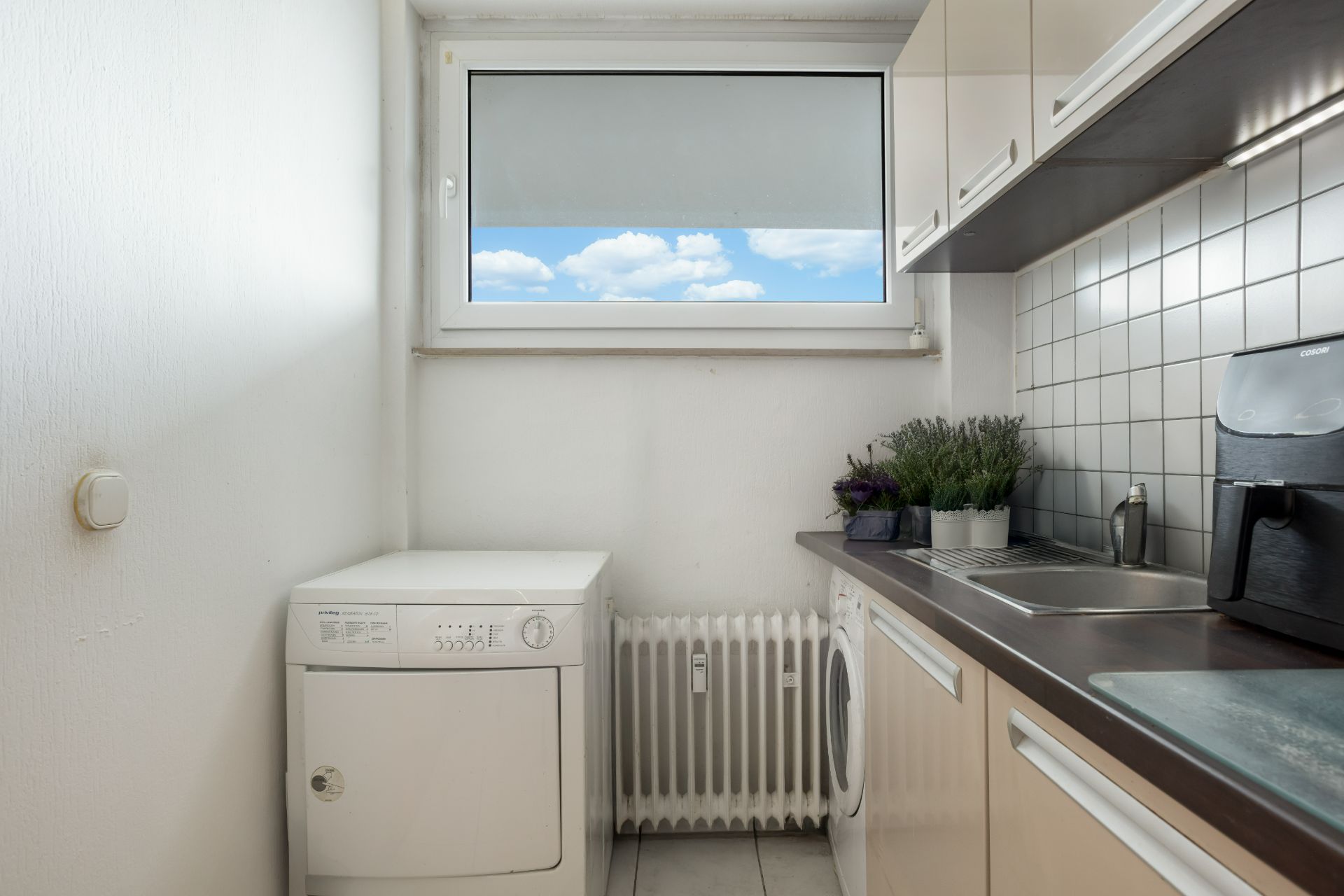 Helle Küche mit Fenster, Waschmaschine, Pflanzen und Kaffeemaschine auf der Arbeitsplatte.