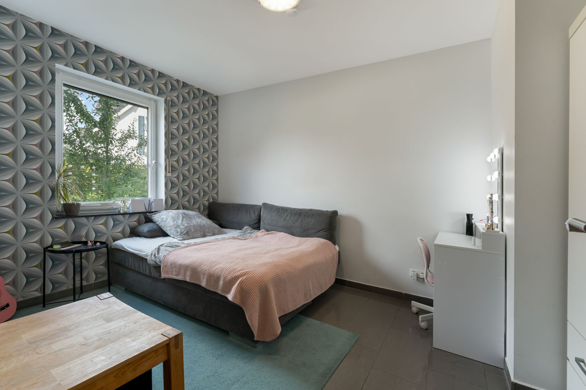Modernes Schlafzimmer mit grauem Bett, Schreibtisch, Fenster und gemusterter Tapete an einer Wand.