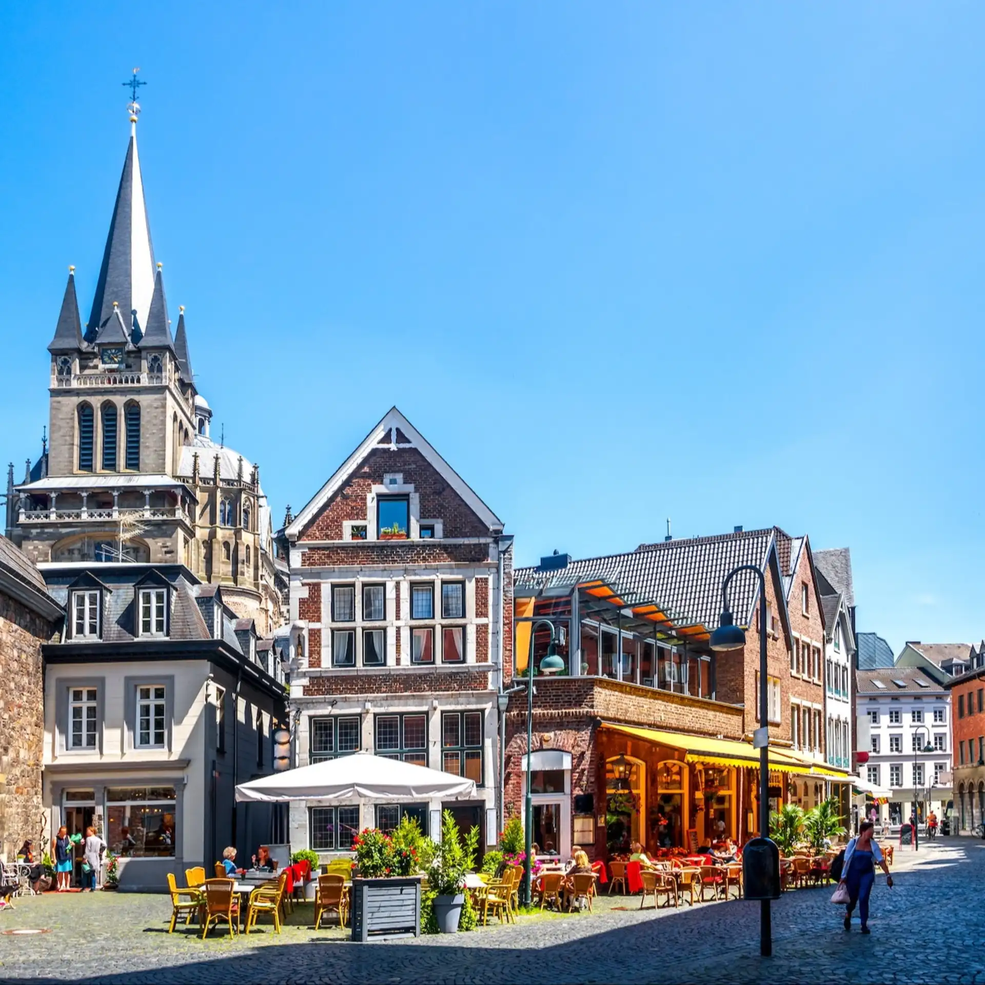 Belebter Platz mit alten Häusern, Straßencafé, Kirchengebäude und Menschen bei Sonnenschein.