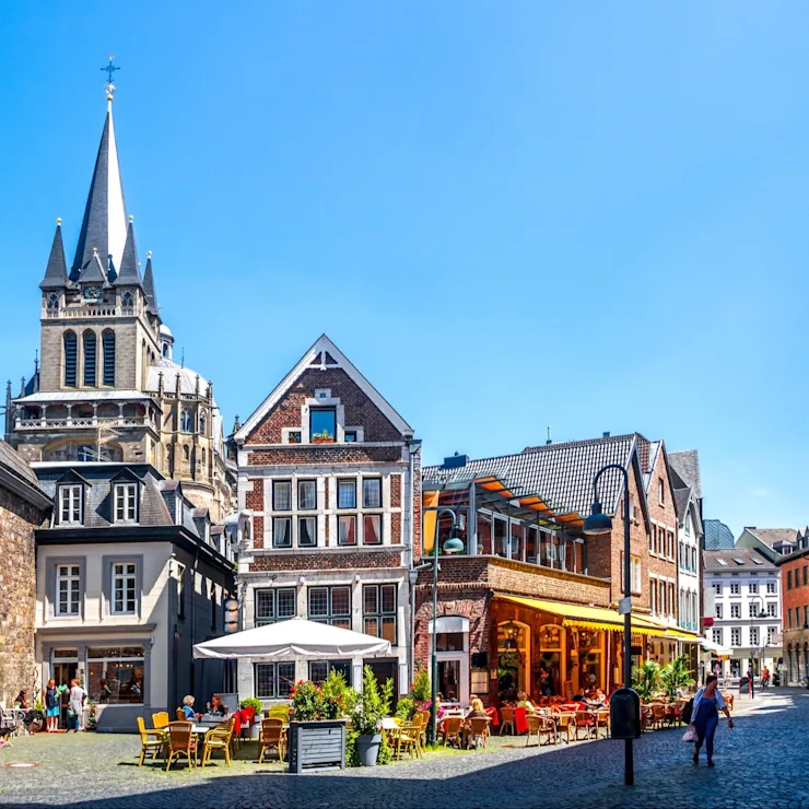 Belebter Platz mit alten Häusern, Straßencafé, Kirchengebäude und Menschen bei Sonnenschein.