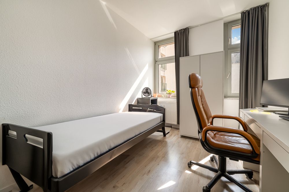 Helles Zimmer mit Einzelbett, Schreibtisch, braunem Bürostuhl und Fenster mit grauen Vorhängen.