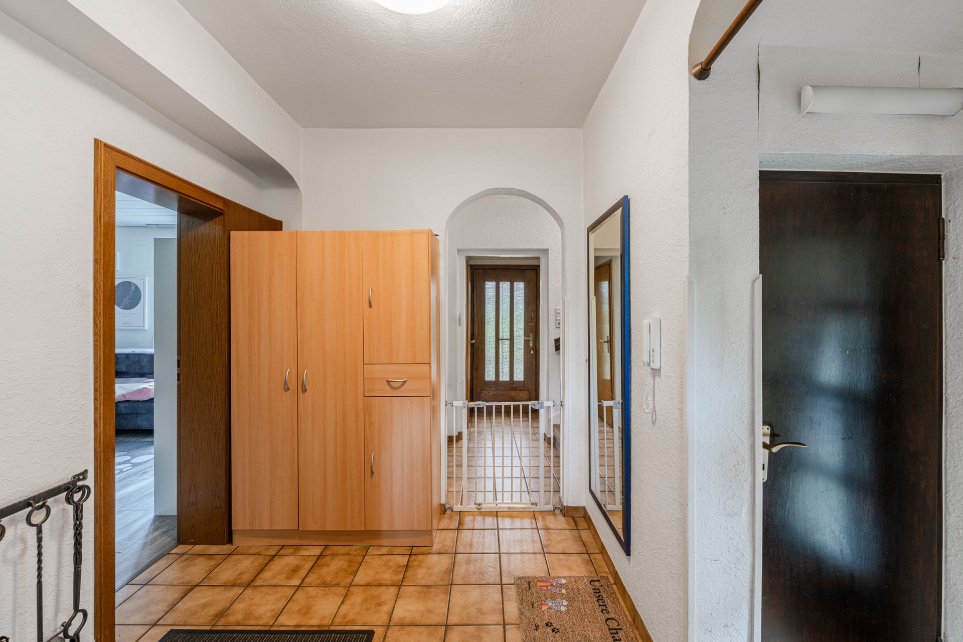 Flur mit Fliesenboden, Holzschrank, Spiegel und Tür, Sicht auf Haustür mit Gitter.