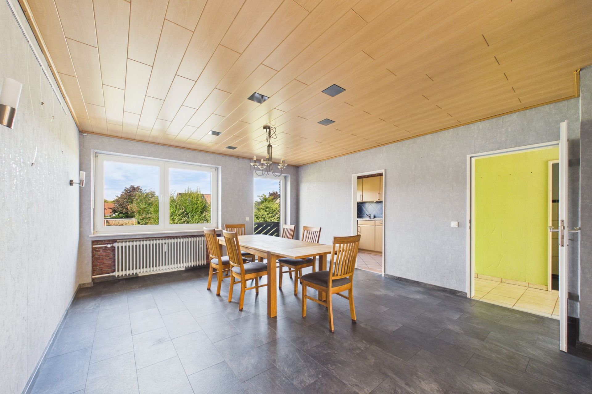 Esszimmer mit Holztisch und sechs Stühlen, großen Fenstern und grauem Fliesenboden.