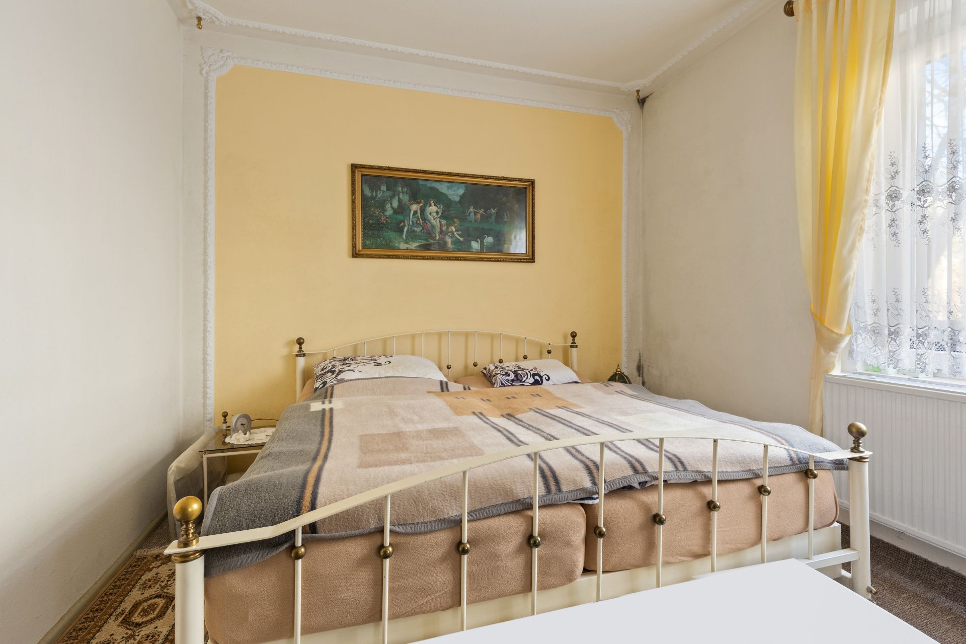 Schlafzimmer mit Doppelbett, gelber Wand, Gemälde und Fenster mit gelben Vorhängen.