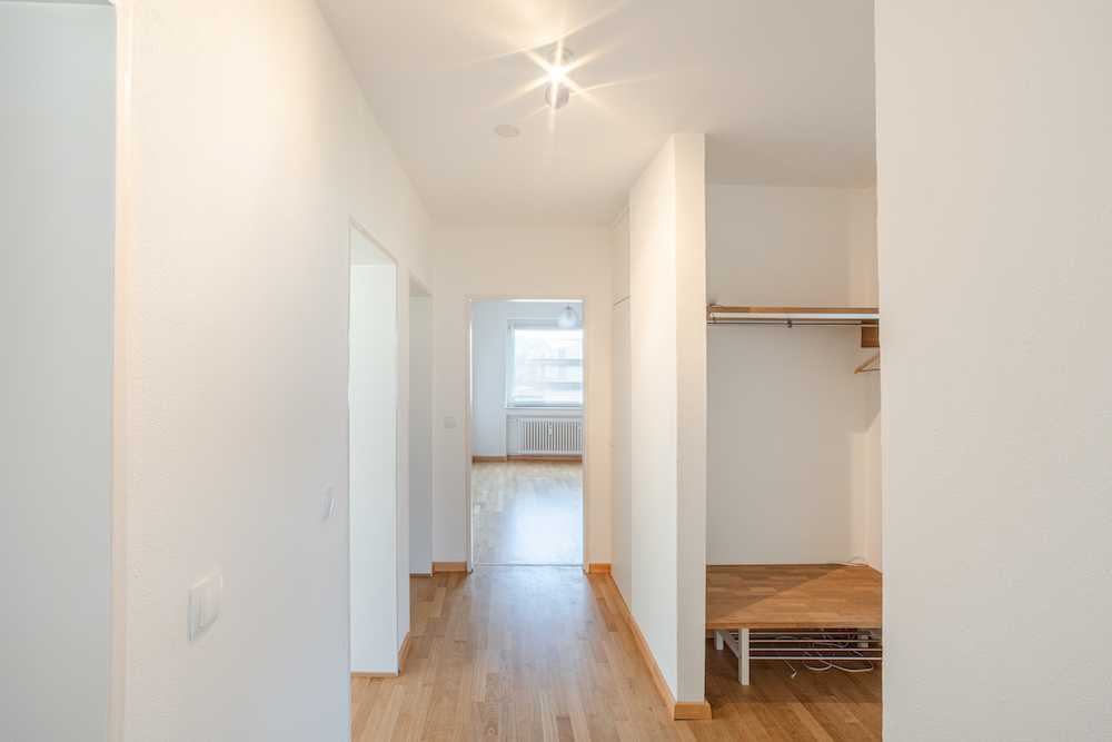 Heller Flur mit Holzfußboden, offenem Einbauschrank und Blick in ein Zimmer mit Fenster.