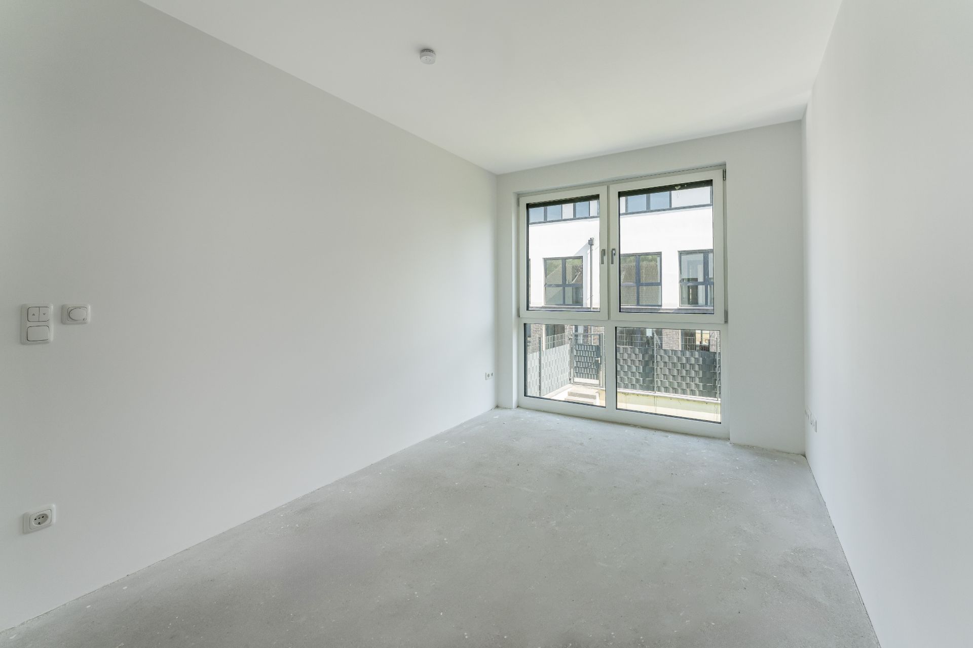 Leerer, heller Raum mit weißen Wänden und großem Fenster, Betonboden.