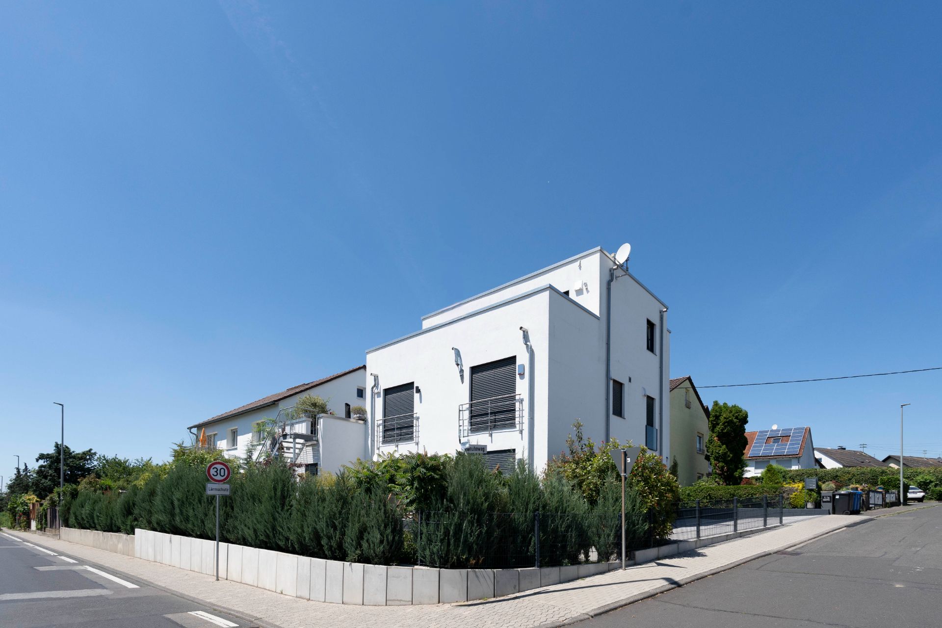 Modernes weißes Wohnhaus mit Rollläden und kleinem Vorgarten an einer Straßenecke.