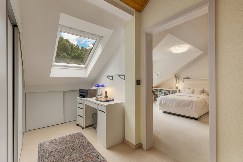 Heller Dachbodenraum mit Schreibtisch, Dachfenster und Blick ins Schlafzimmer mit Doppelbett.