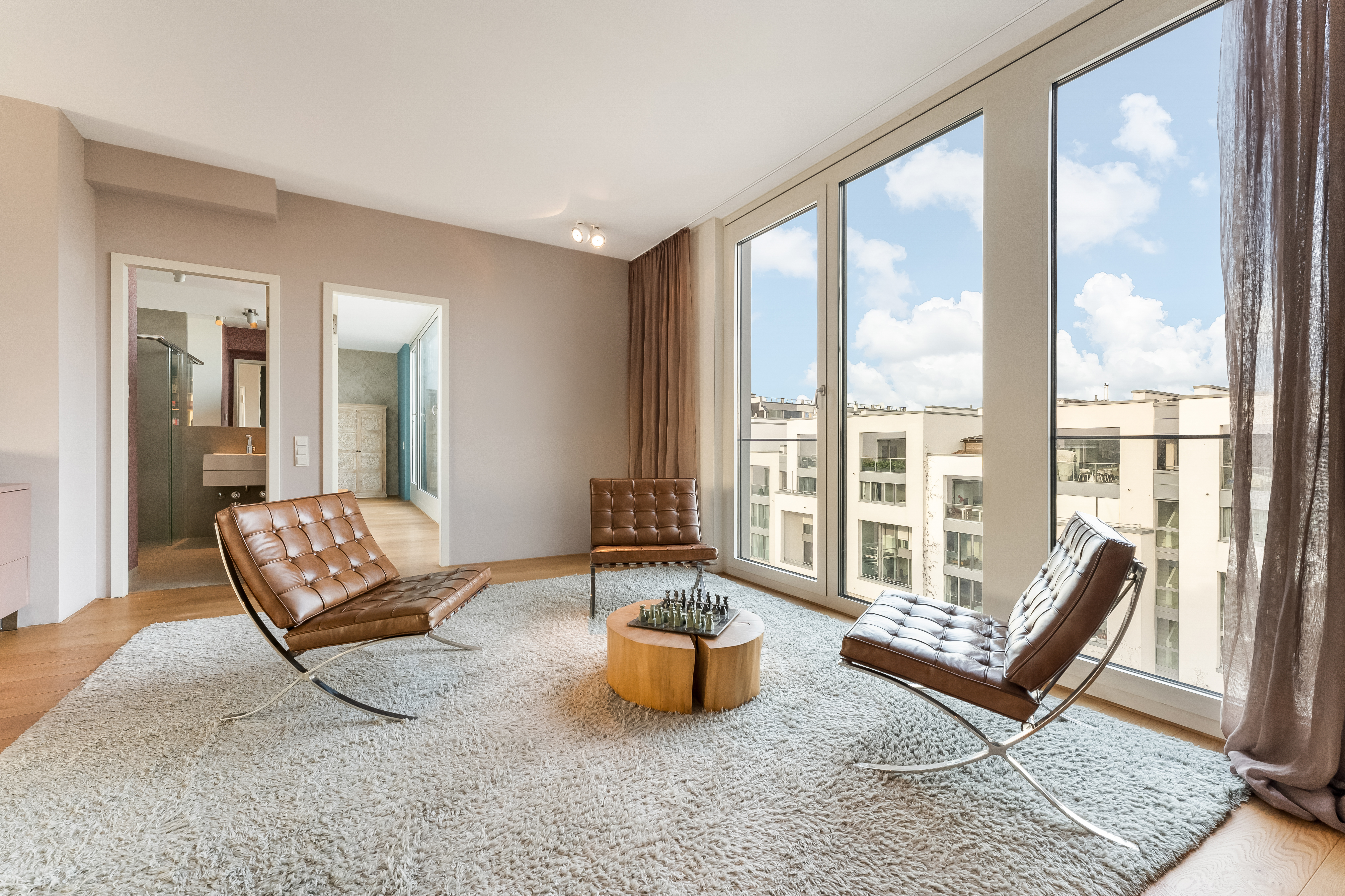 Modernes Wohnzimmer mit drei braunen Sesseln, rundem Tisch und großen Fenstern mit Stadtblick.