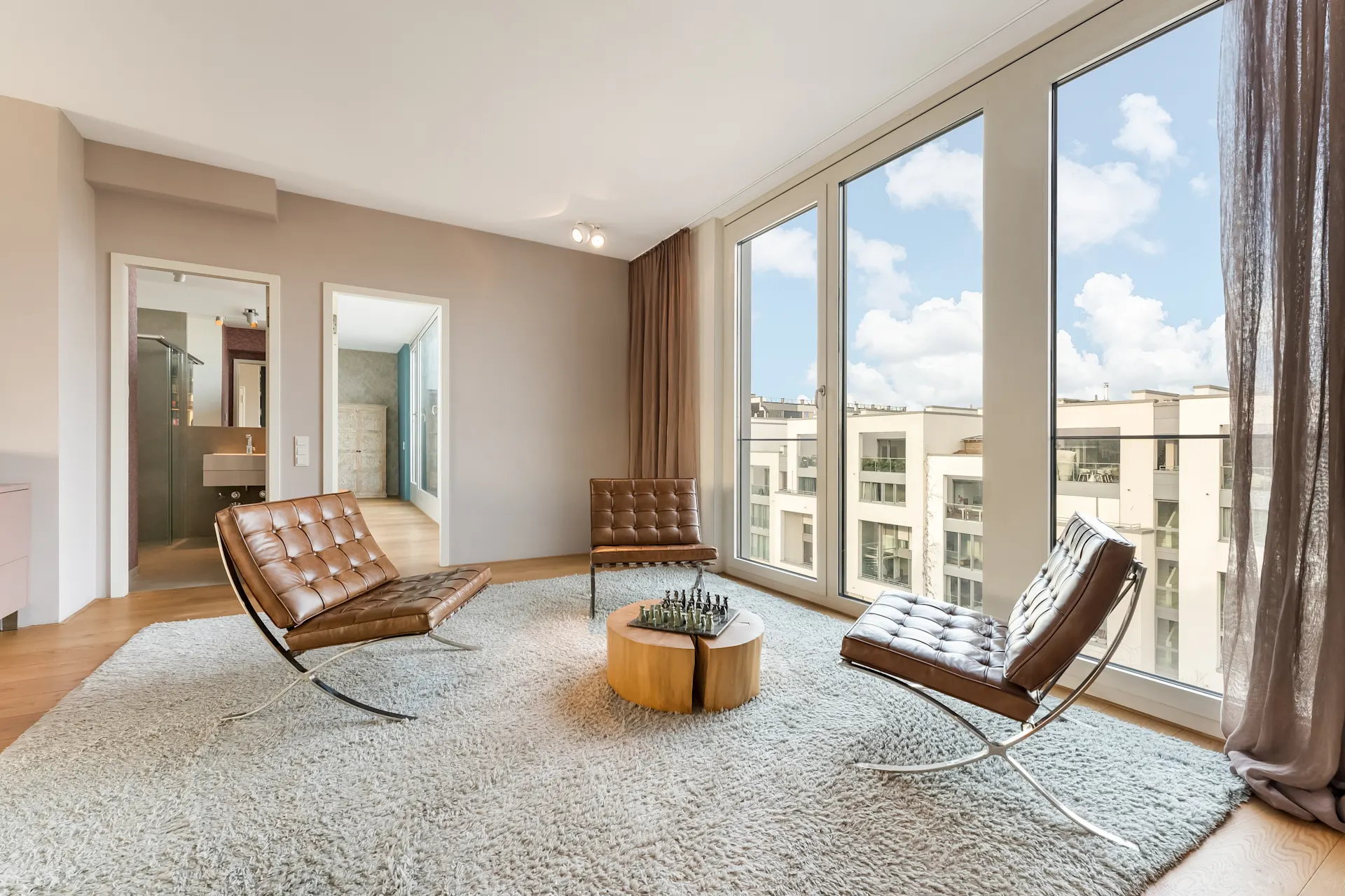 Modernes Wohnzimmer mit drei braunen Sesseln, rundem Tisch und großen Fenstern mit Stadtblick.