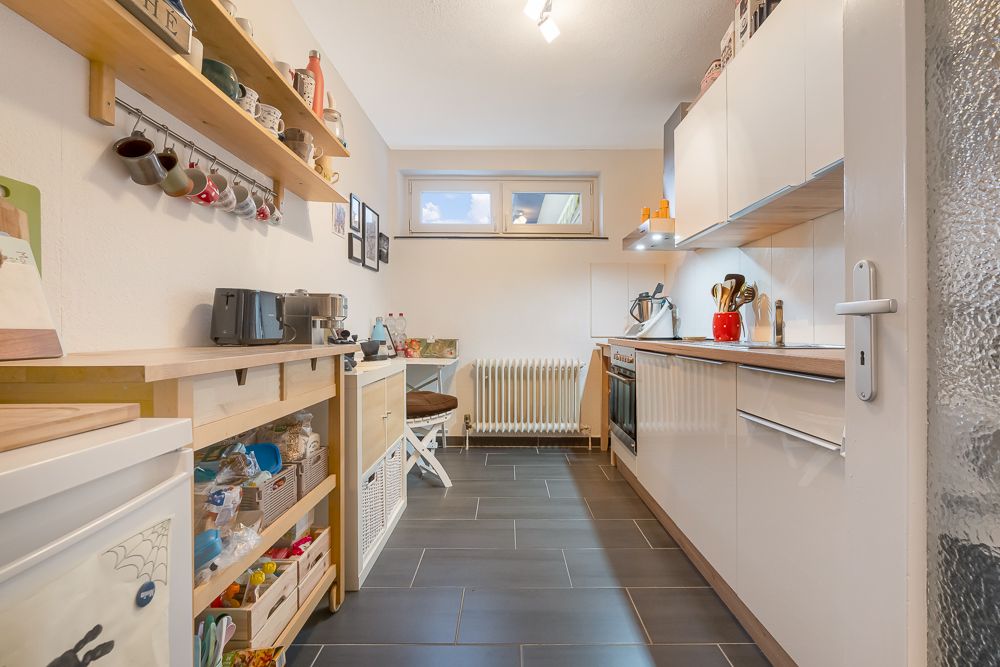 Helle, schmale Küche mit weißen Schränken, Holzregalen und dunklem Fliesenboden.