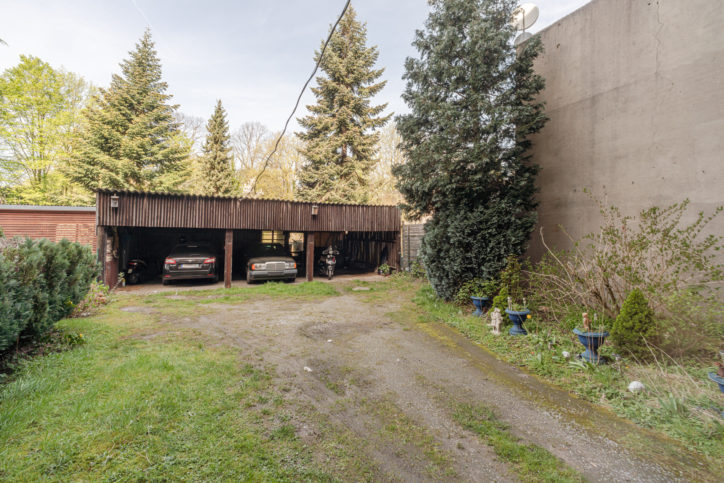 Offener Carport mit zwei Autos und einem Motorrad, umgeben von Bäumen und Pflanzen.