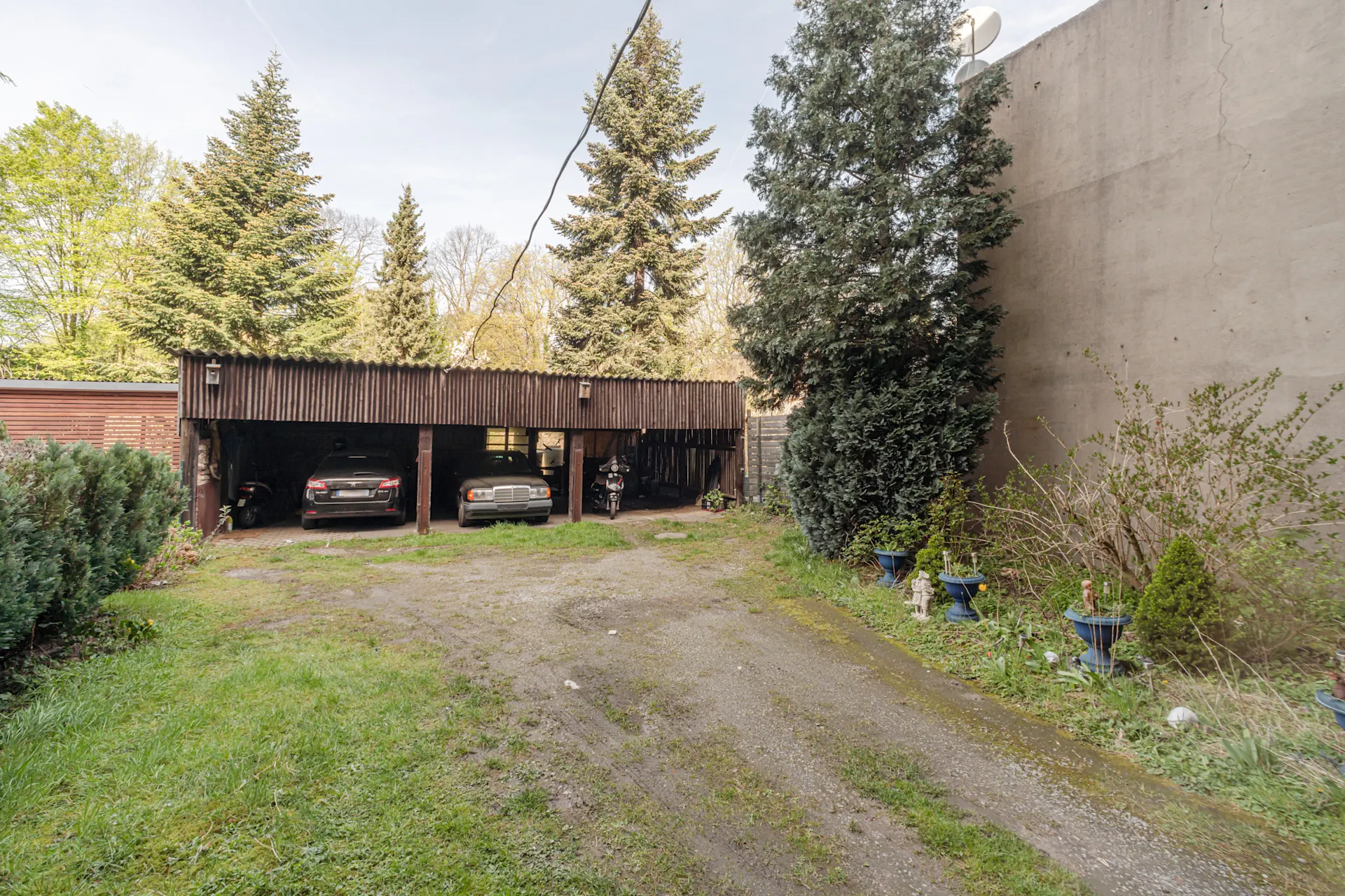 Offener Carport mit zwei Autos und einem Motorrad, umgeben von Bäumen und Pflanzen.