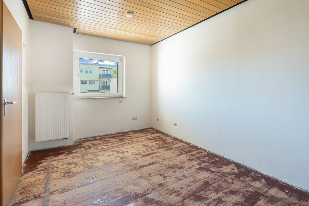 Leerer Raum mit Fenster, Holzdecke, weiß gestrichenen Wänden und abgenutztem Boden.