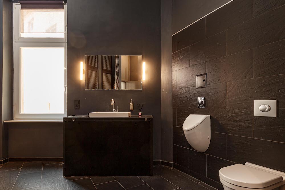 Modernes Badezimmer mit Fenster, Spiegel, Waschbecken, Urinal und Toilette, dunkle Fliesenwände.