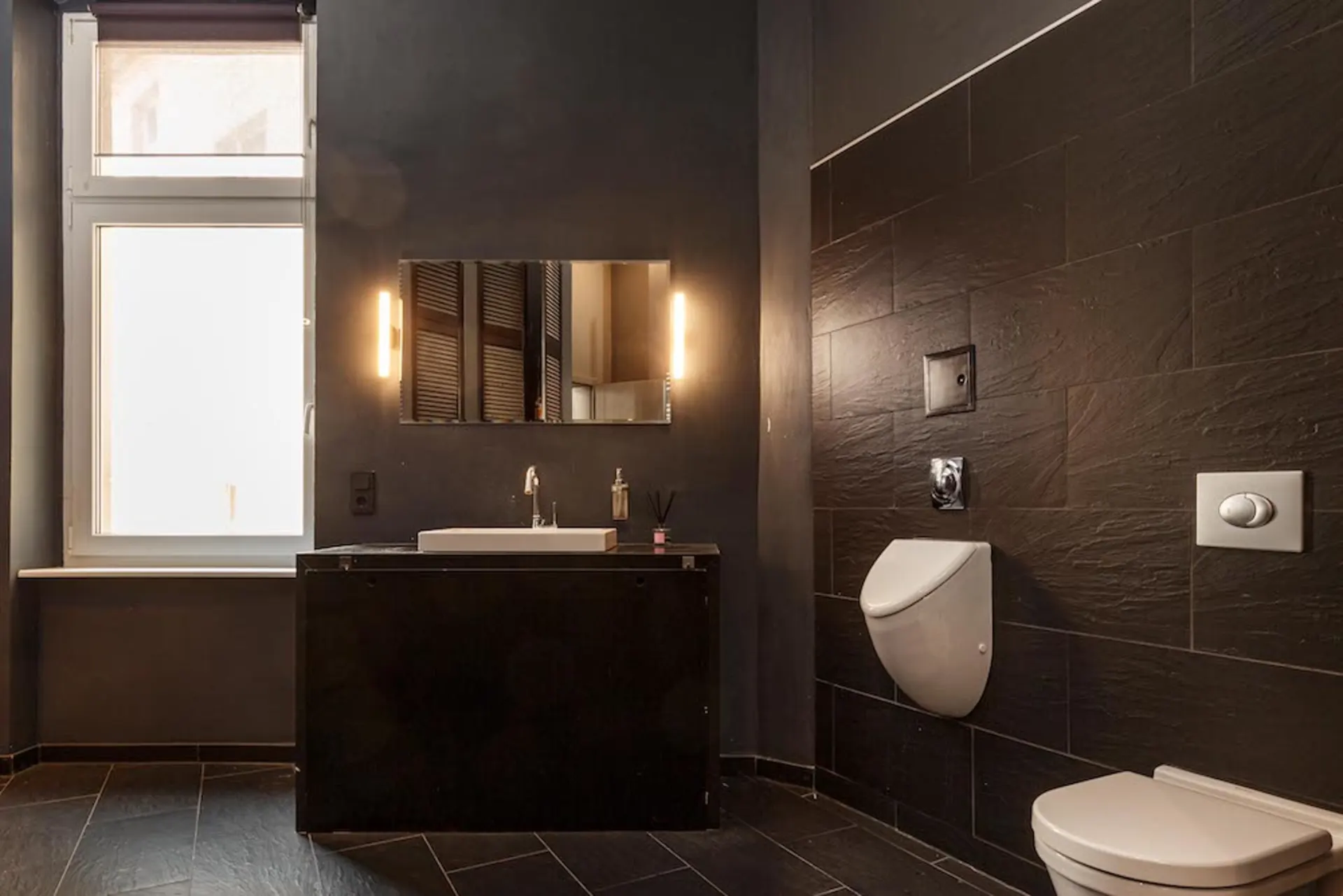 Modernes Badezimmer mit Fenster, Spiegel, Waschbecken, Urinal und Toilette, dunkle Fliesenwände.