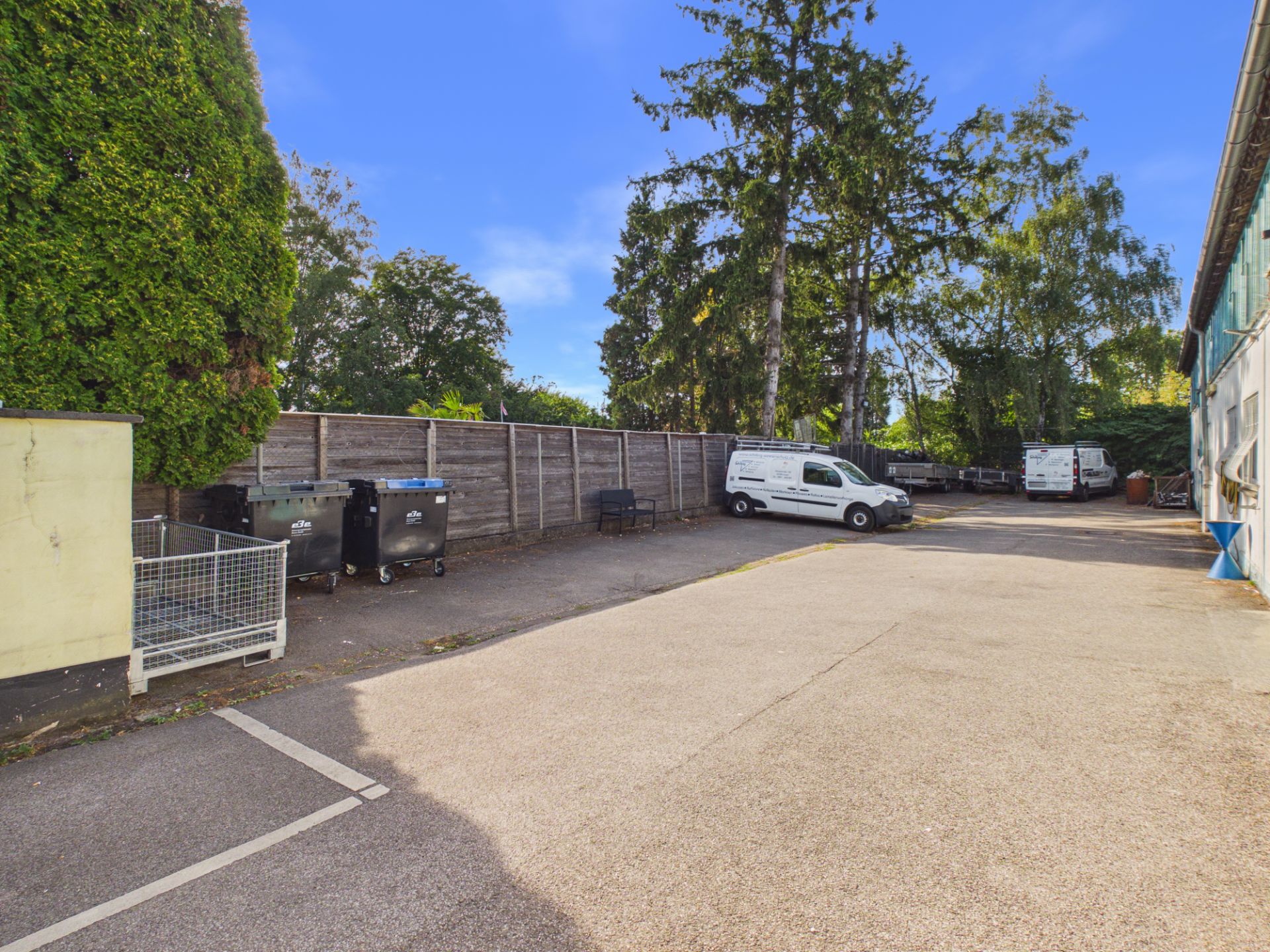 Gepflasterter Parkplatz mit Mülltonnen, Transportern und Bäumen im Hintergrund.