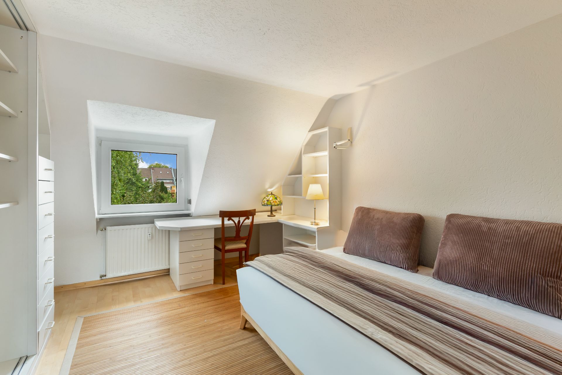 Helles Schlafzimmer mit Bett, Schreibtisch am Fenster und Regalwand.