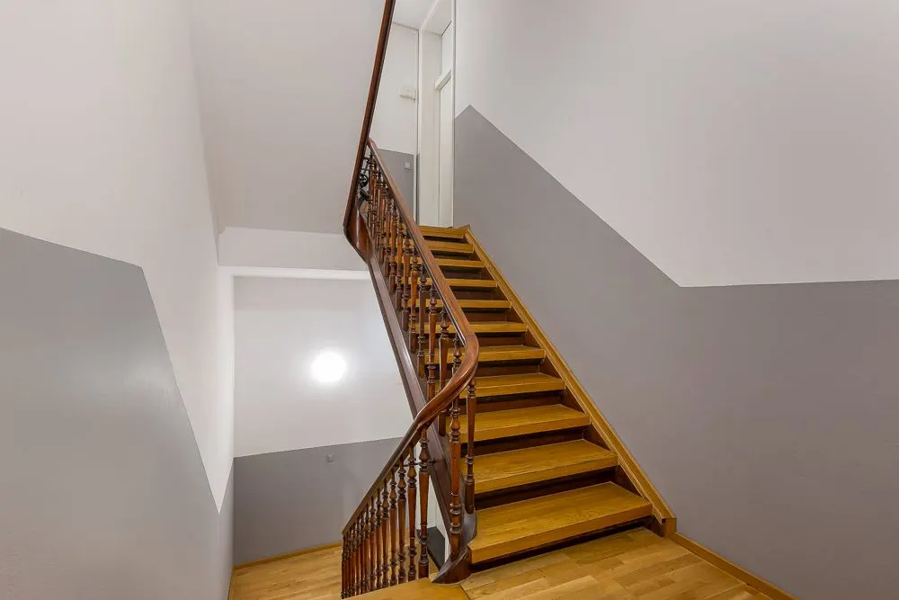 Holztreppe mit Holzgeländer in einem Hausflur mit grauen und weißen Wänden.