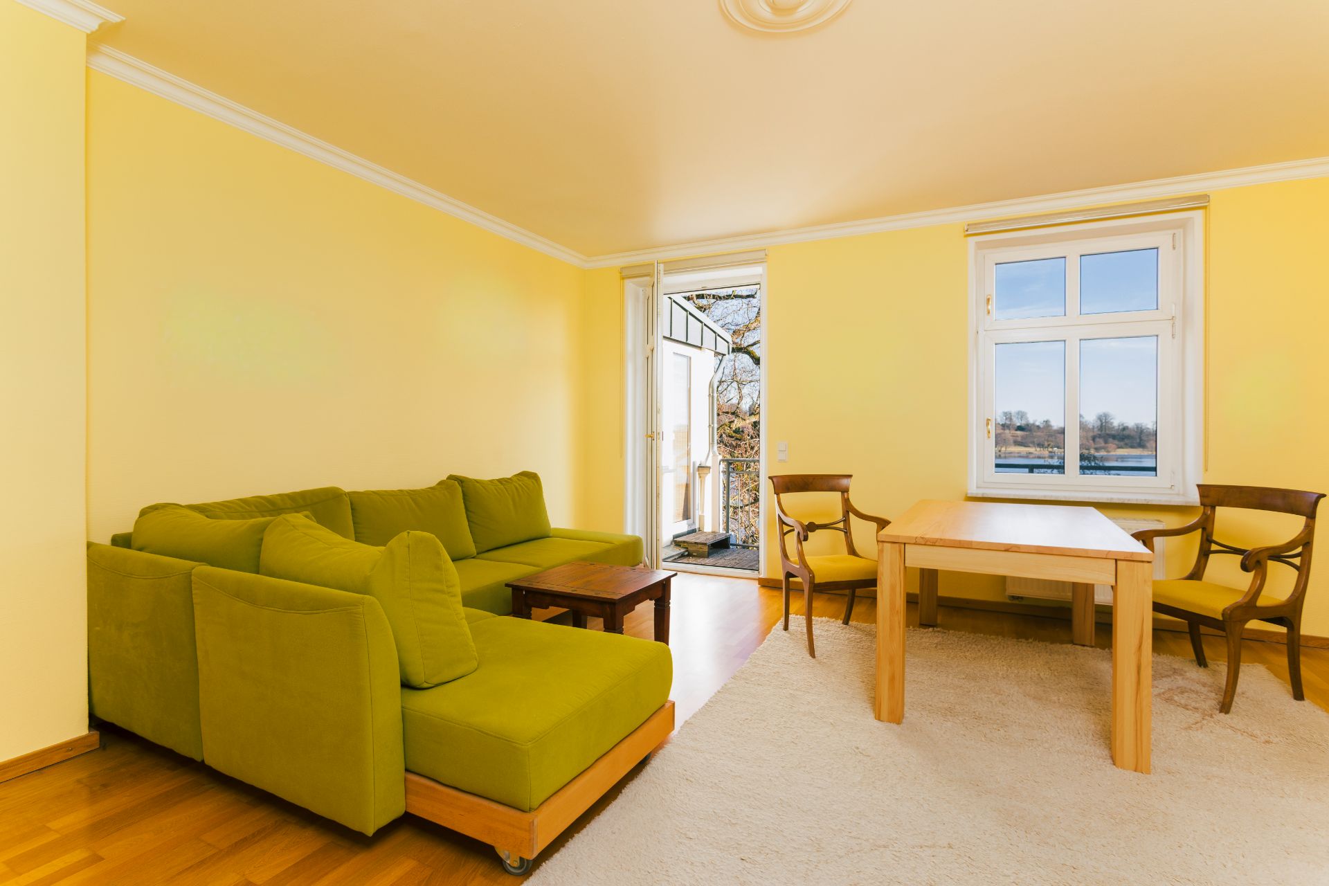 Wohnzimmer mit grünem Sofa, Holztisch, zwei Stühlen, Teppich und Fenstertür zum Balkon.