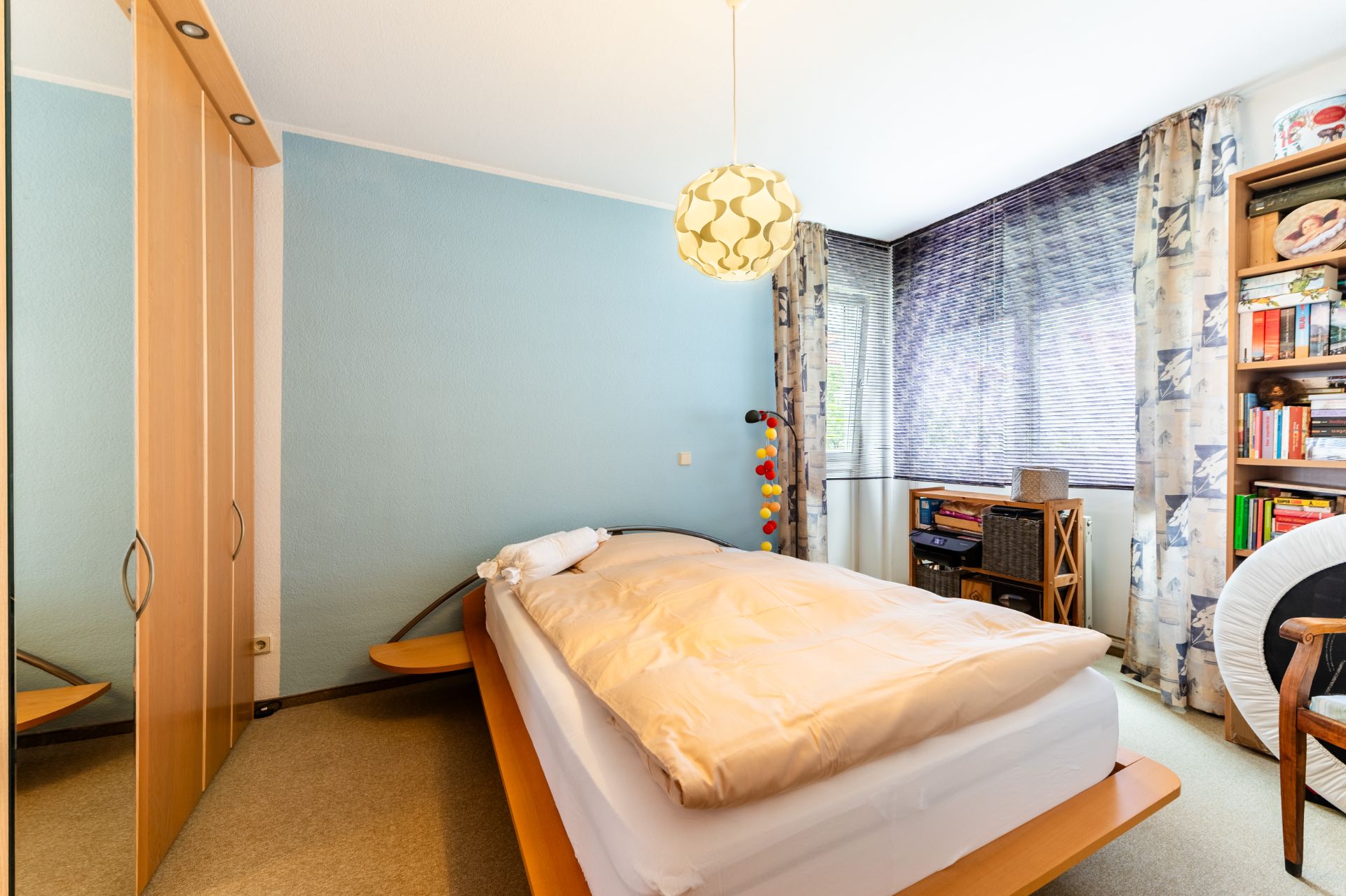 Helles Schlafzimmer mit Doppelbett, Kleiderschrank, Regal und einem großen Fenster mit Vorhängen.